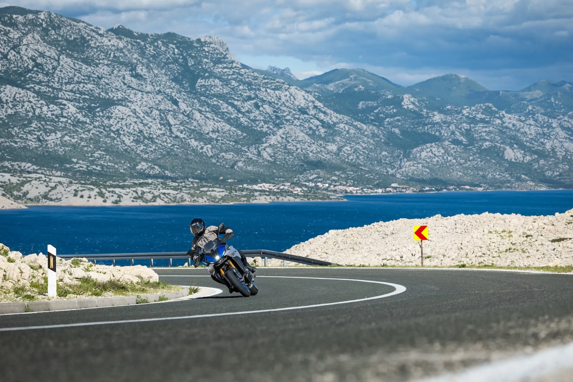 NastyNils riding a blue Suzuki GSX-S1000GX sport-tourer through a sweeping left-hand corner on a coastal mountain road, likely along the Croatian Adriatic. The bike leans into the bend with the rider in full gear and a touring windscreen raised. A red-and-yellow chevron curve-warning sign marks the apex on the right shoulder. Steep limestone mountains rise behind a deep-blue stretch of sea, the sky overcast with broken clouds. Daylight, dry asphalt.