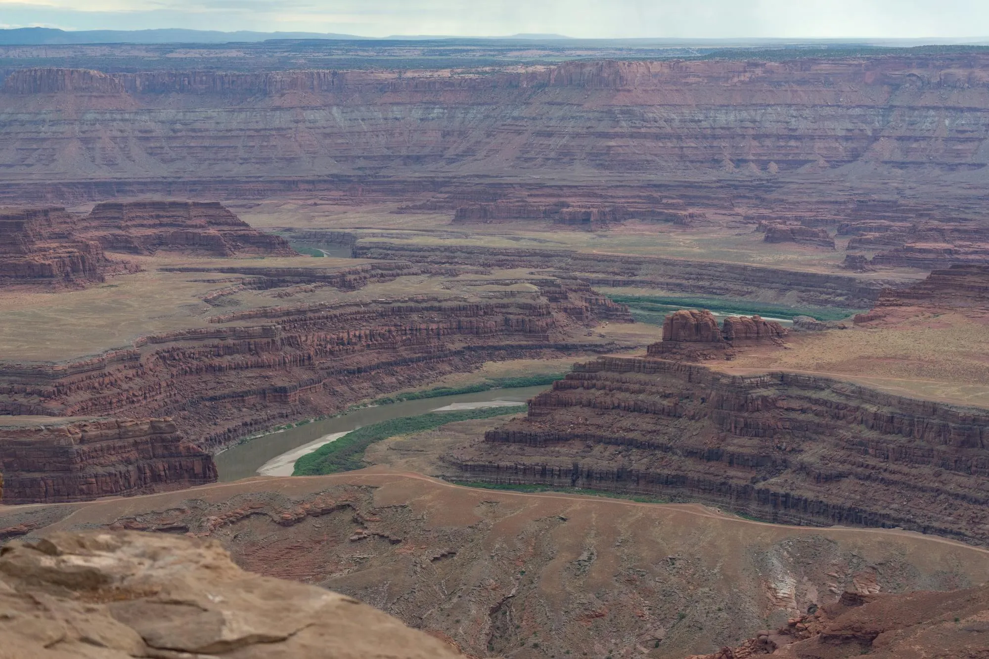 Aerial panoramic view of Dead Horse Point State Park near Moab, Utah. The Colorado River winds through deeply layered red-rock canyons and mesas characteristic of the high desert terrain. The arid landscape extends toward distant mountains under a partly cloudy sky. No motorcycle or rider visible. Daylight conditions, good visibility. Stock photograph by Drew Burks from Pexels.