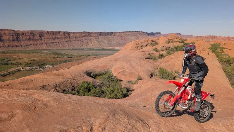 NastyNils leaning a red Beta RR 2T X-Pro 200 through a curving trail section above a Utah river valley with distant red-rock buttes on the horizon.