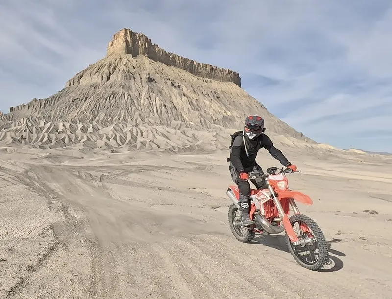 NastyNils standing on the pegs of a red Beta RR 2T X-Pro 200 through loose sand in front of a dramatic layered rock formation in the Utah desert.