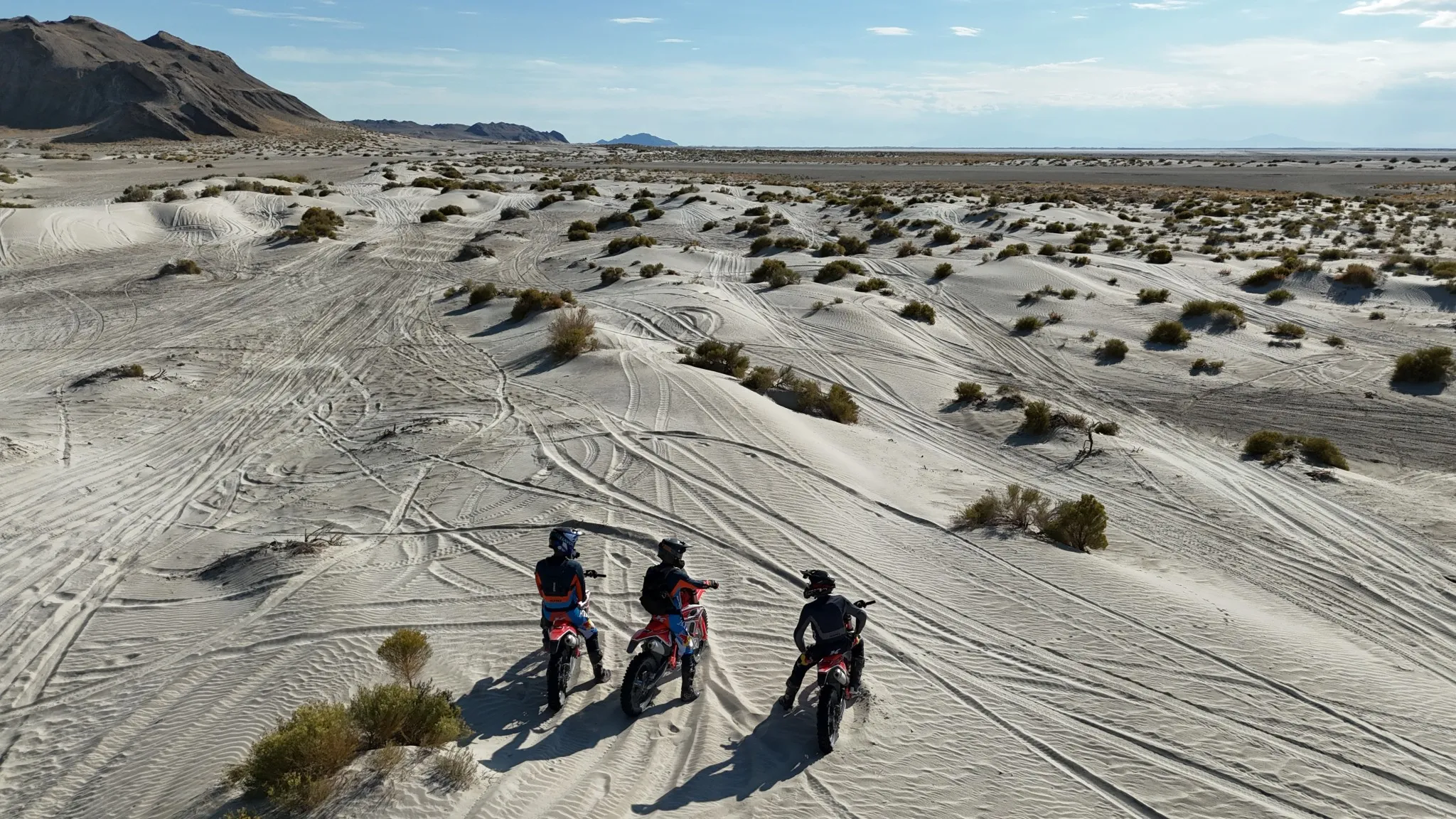 Three off-road riders standing with their dirt bikes in a Utah desert landscape of sand dunes, sparse vegetation, and visible tire tracks, mountains in the background.