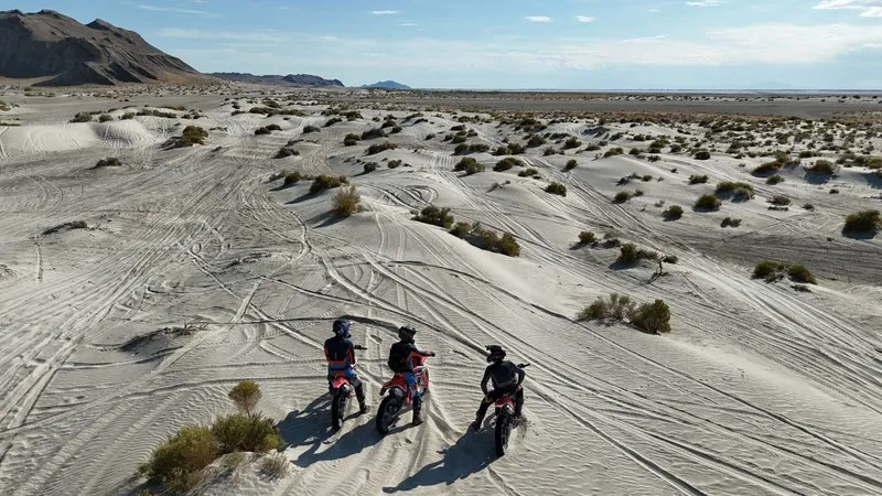 Three off-road riders standing with their dirt bikes in a Utah desert landscape of sand dunes, sparse vegetation, and visible tire tracks, mountains in the background.
