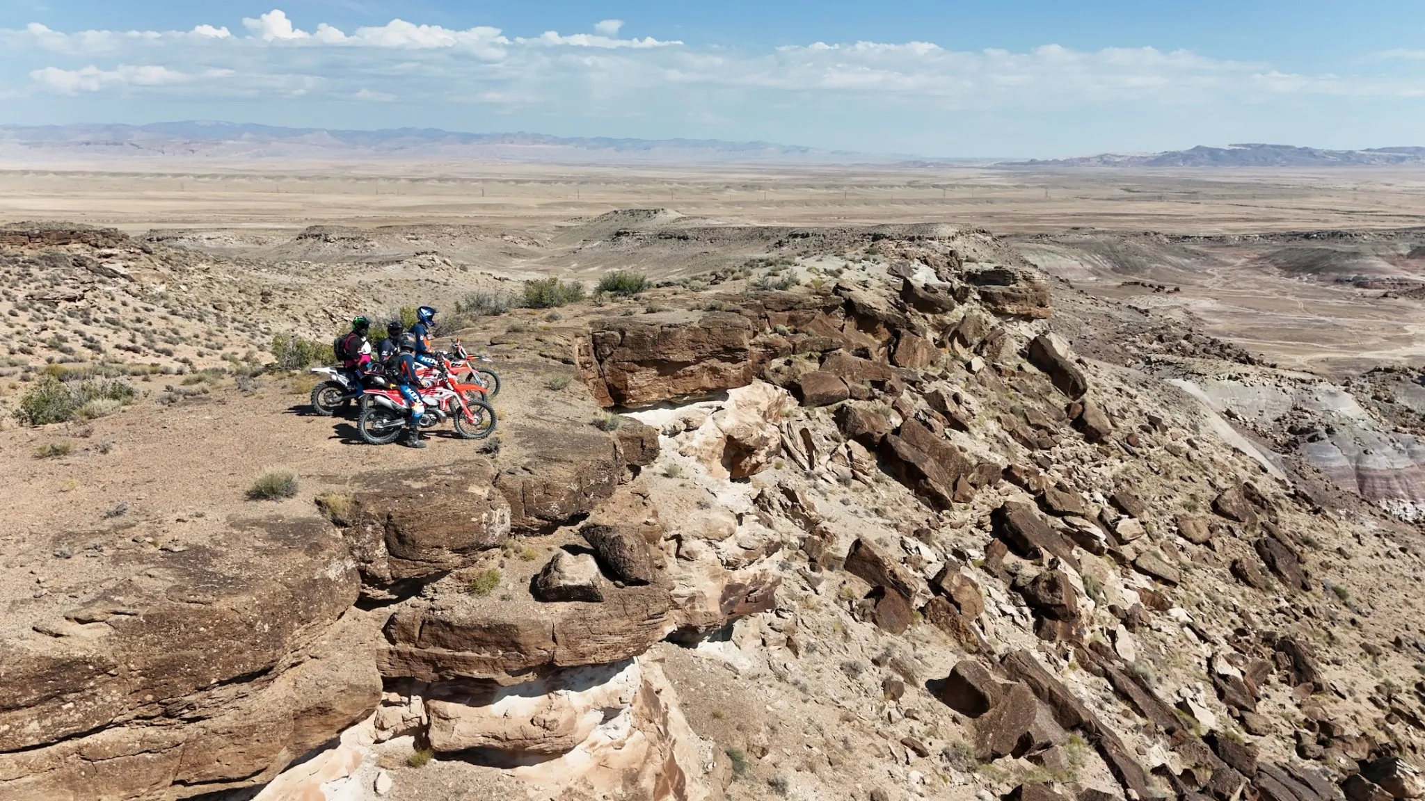 Two riders stopped on a rocky sandstone ridge overlooking the vast Utah desert plain, sparse vegetation extending to distant mountain ranges under a partly cloudy sky.
