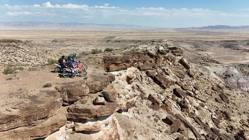 Two riders stopped on a rocky sandstone ridge overlooking the vast Utah desert plain, sparse vegetation extending to distant mountain ranges under a partly cloudy sky.