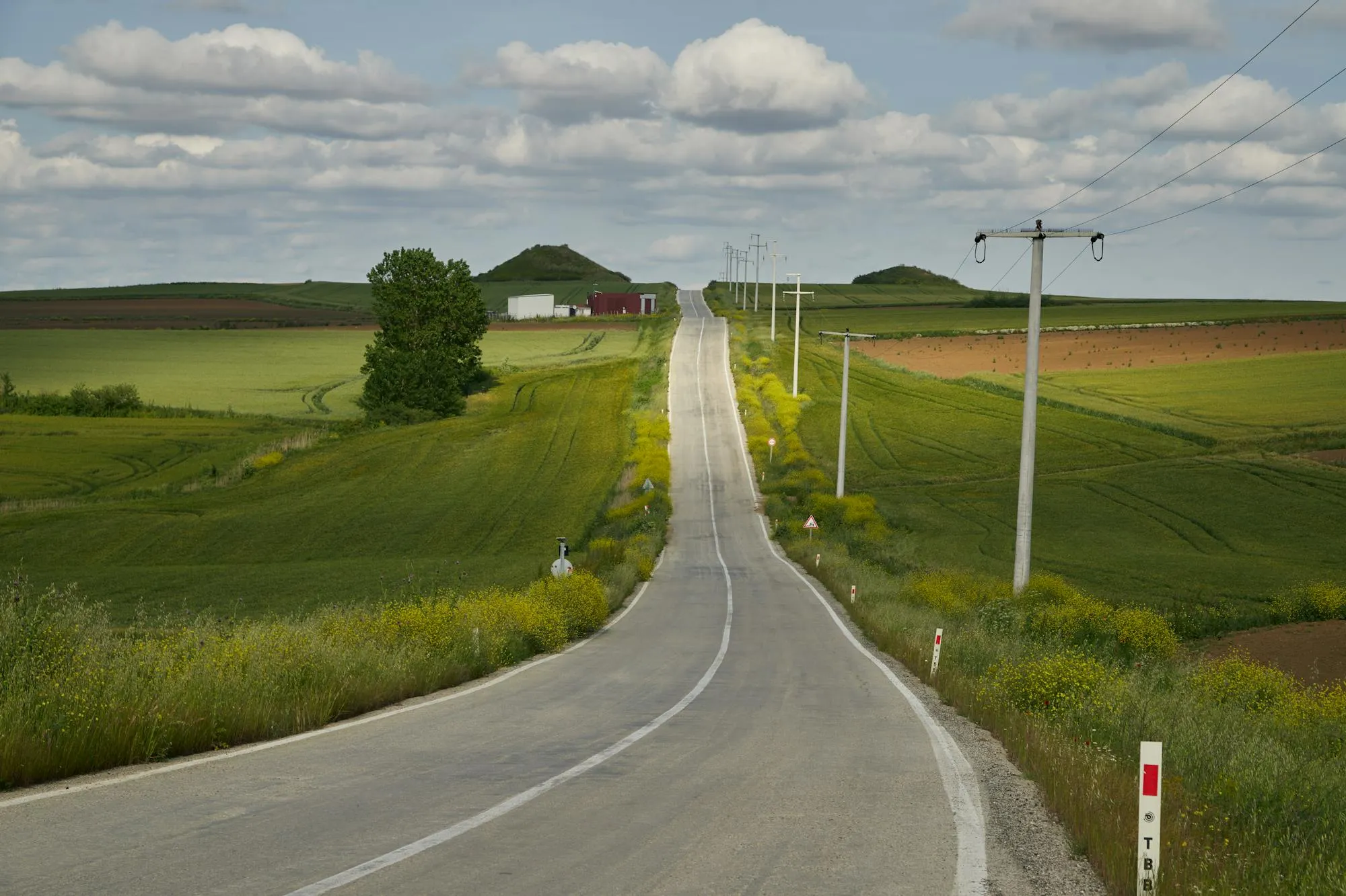 A long, straight paved road stretches toward the horizon through agricultural fields in Vermont. Power lines and a mailbox are visible along the roadside. Lush green vegetation, cumulus clouds, good daylight conditions. The landscape is flat to gently rolling. Asphalt pavement appears to be in good condition. Rural American setting.