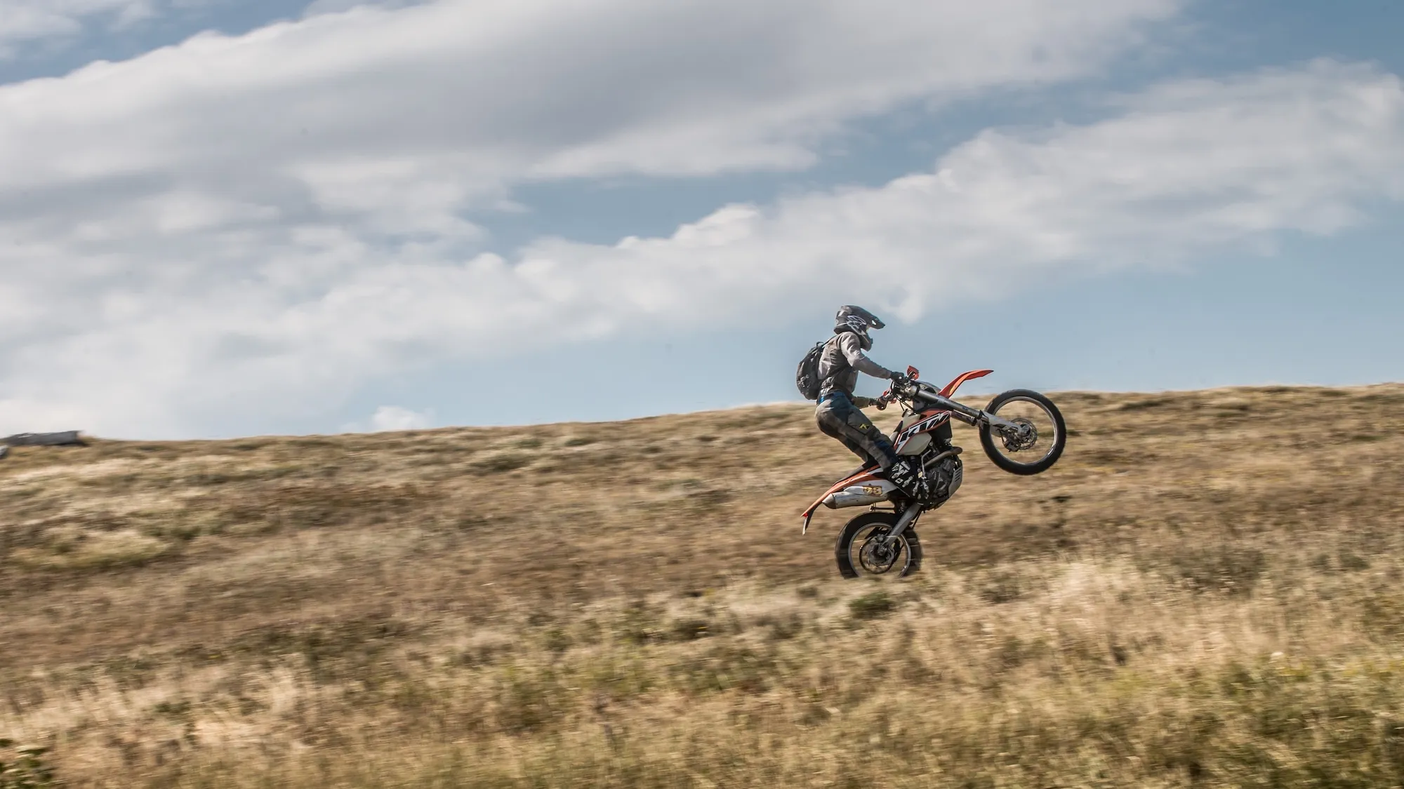 NastyNils riding a KTM enduro motorcycle on a dry, grassy hillside, performing a wheelie with the front wheel lifted high. He wears full off-road gear including a full-face helmet, body armor with hydration pack, and protective pants. The bike shows the orange-and-white KTM livery typical of their enduro lineup. Open landscape with rolling hills and a cloudy blue sky in the background. Daylight, dry conditions, motion blur in the foreground grass indicates speed.